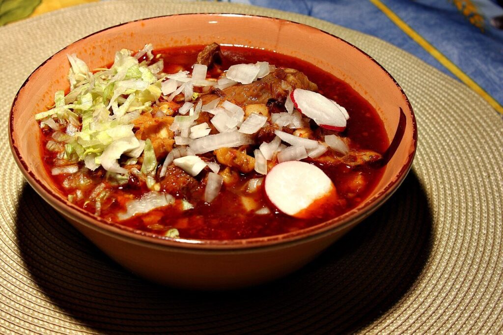 Bowl of red pozole with pork and hominy topped with shredded cabbage, radish, and onion