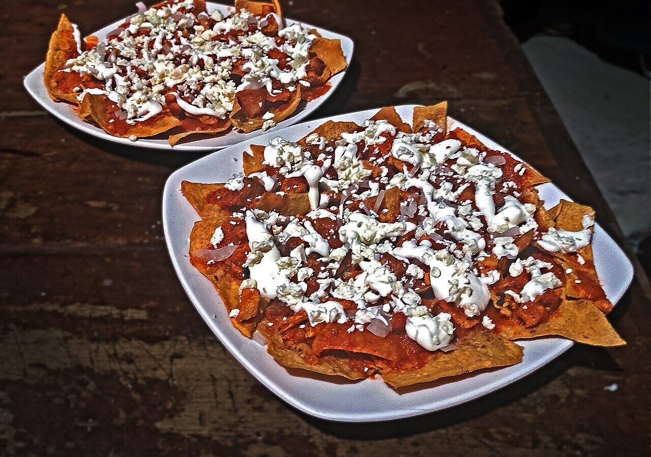 Red chilaquiles topped with crema, crumbled cheese, and onion on white plates